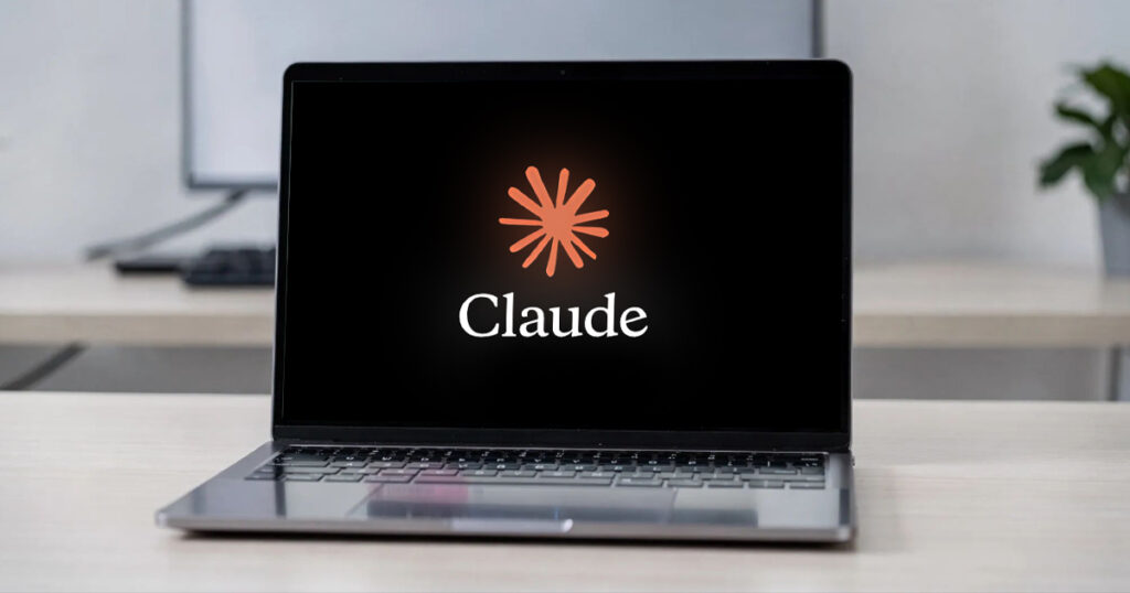 Open laptop on a light wood desk displaying the Claude logo, an orange starburst above the word "Claude" on a black screen, in a bright modern office.