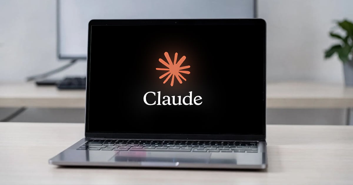 Open laptop on a light wood desk displaying the Claude logo, an orange starburst above the word "Claude" on a black screen, in a bright modern office.
