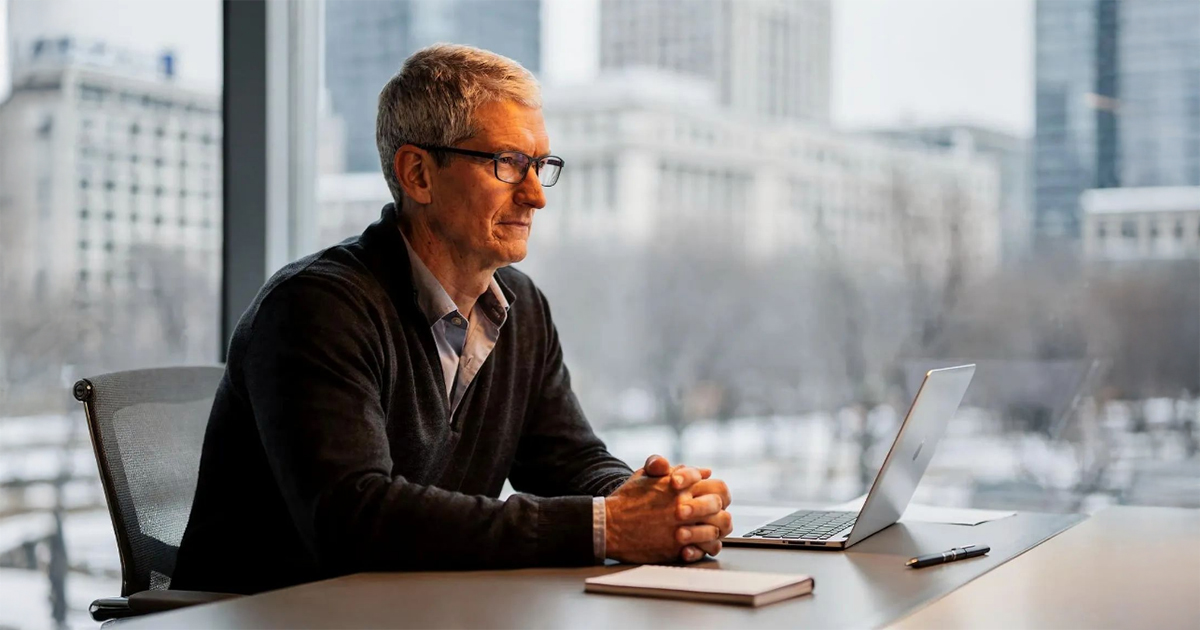 A man wearing glasses sits at a desk with a laptop, hands clasped, looking thoughtfully out a window in an office setting.