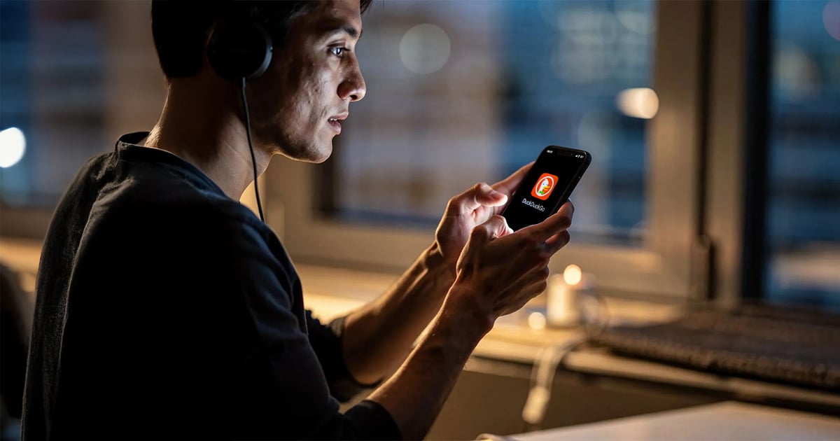 A side-profile shot of a young man wearing headphones, looking intently at his smartphone screen which displays the DuckDuckGo logo. He is in a dimly lit indoor setting at night, with a blurred window and a small candle in the background.