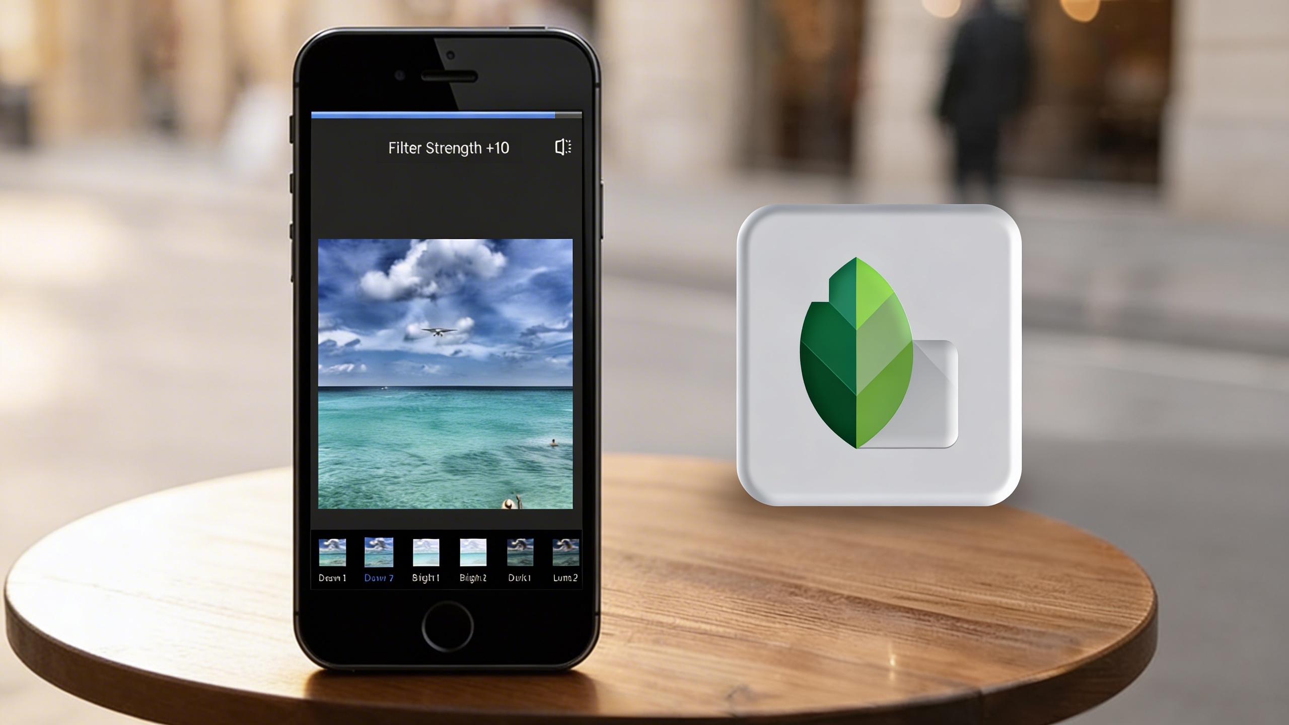 Smartphone displaying a photo editing app with filter options and an ocean image, placed on a wooden table beside the Snapseed logo icon.