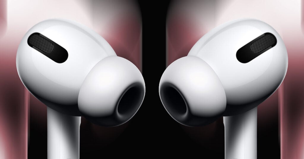 Extreme close-up of a pair of white AirPods Pro facing each other, silicone ear tips and mesh microphone vents visible against a dark background with soft red and pink lighting.