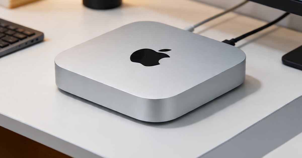 Silver Mac mini with a black Apple logo on top, positioned on a white desk beside a keyboard, monitor stand, and cables.