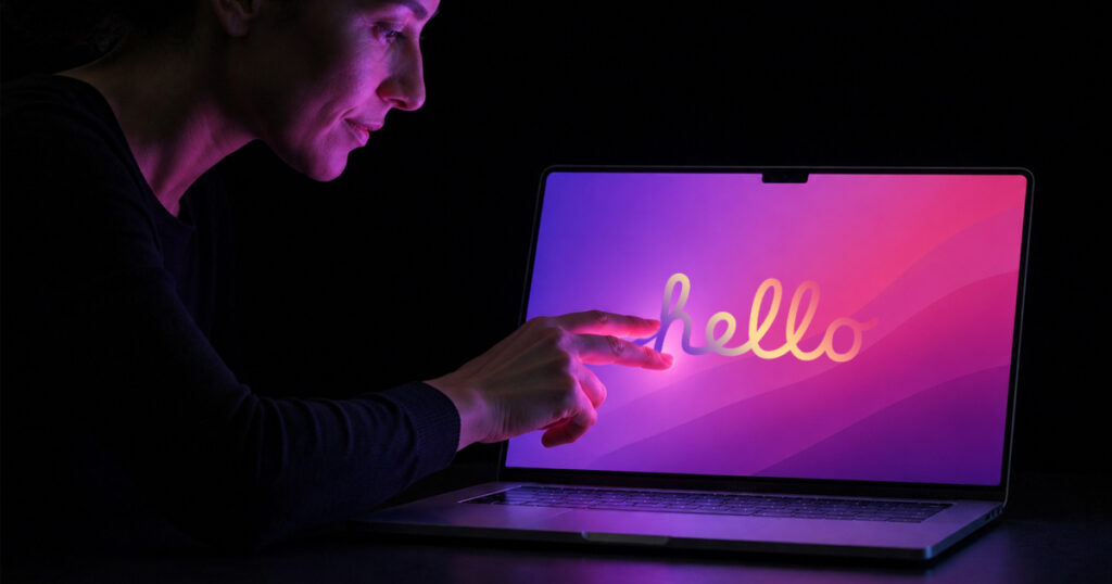 A finger taps a radial control menu on a silver MacBook Pro's touchscreen display showing a colorful abstract swirl wallpaper and a Dynamic Island-style media player notch, sitting on a wooden cafe table.
